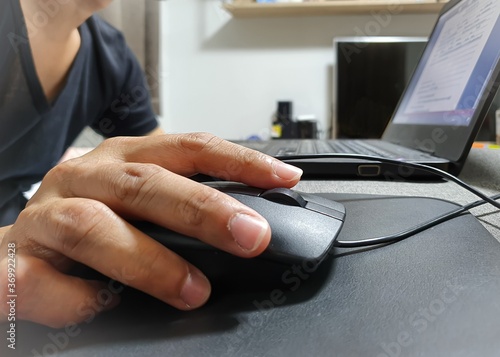 Asian man's hand is holding black mouse while working on laptop at home. Work from home concept for covid-19 prevention and control. Selective focus, blur background.