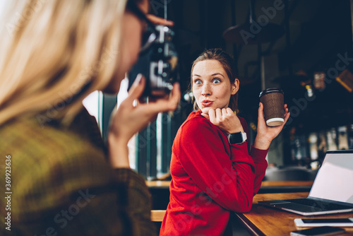 Positive female blogger with funny grimace on face posing on vintage camera having fun during coffee break.Hipster girl focusing to make photo of best friend for publication in social networks