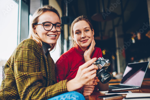Half length portrait of happy skilled female amateur smiling at camera together with positive best friend resting in stylish coffee shop.Cheerful two students with vintage camera sitting in coworking