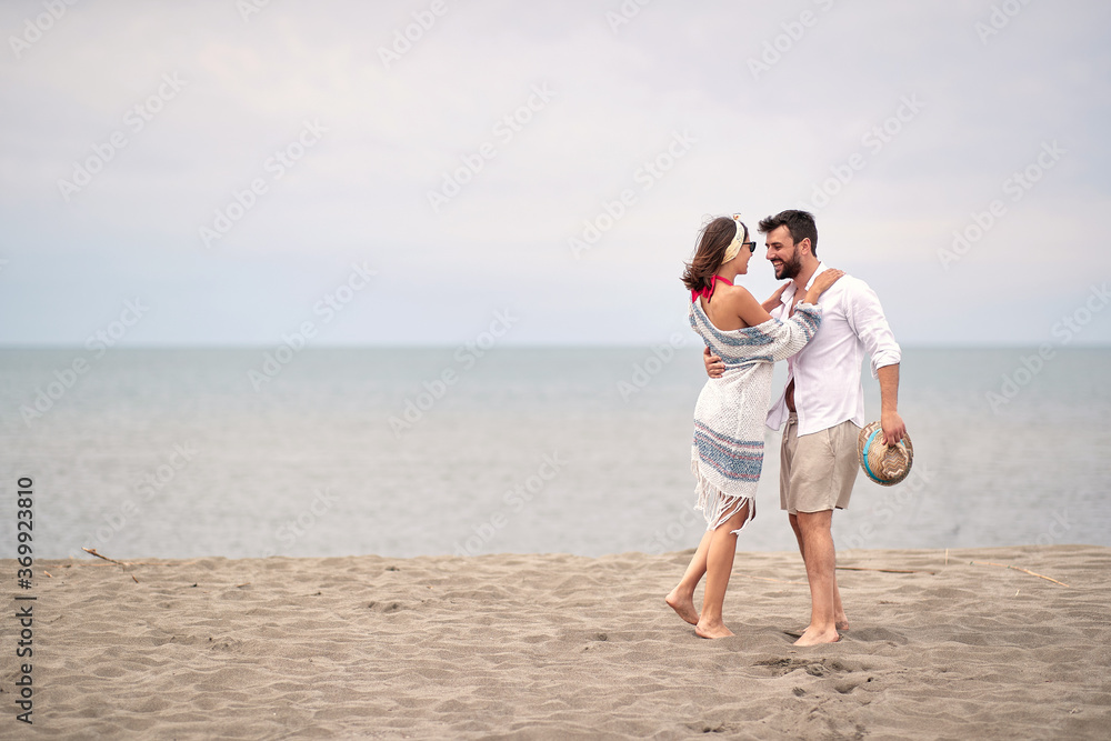 young adult couple hugging, looking each other, standing barefoot at sandy beach