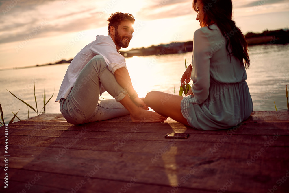 young beautiful caucasian couple sitting on wooden platform by the lake, looking each other, talking