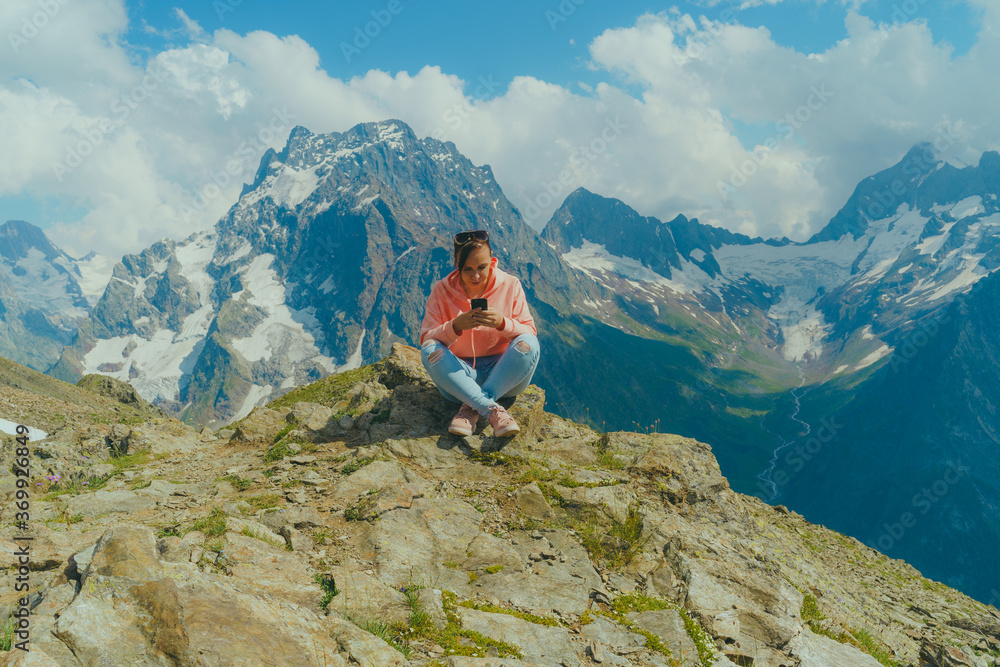 Female traveler using smartphone in mountains. Full body woman sitting on rock and browsing smartphone against cloudy sky on sunny day in mountains