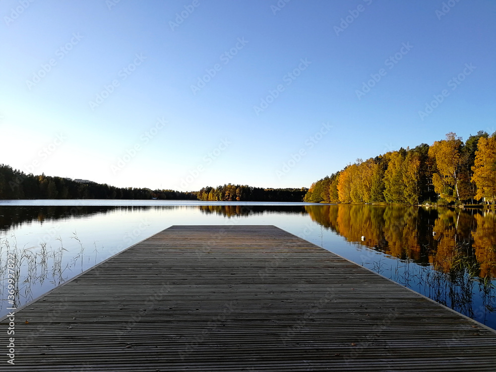 Naklejka premium Colorful autumn landscape. A pier on the calm water of the lake. Yellow and red trees in the rays of the setting sun.