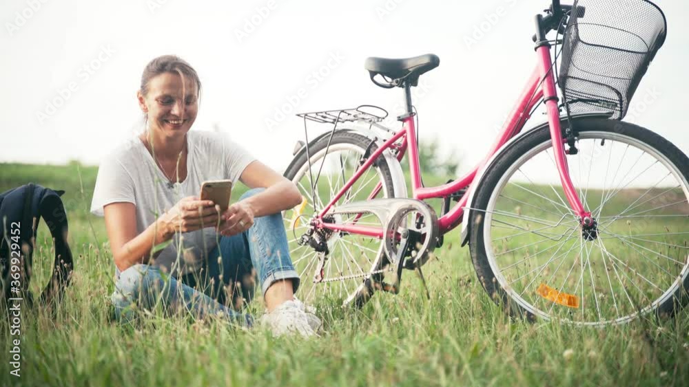 A young cheerful woman sits in the grass on the field with a smartphone next to her backpack and a bicycle with a basket and enjoys the fresh air. A young cheerful woman sits in the grass on the field with a smartphone next to her backpack and a bicycle with a basket and enjoys the fresh air.