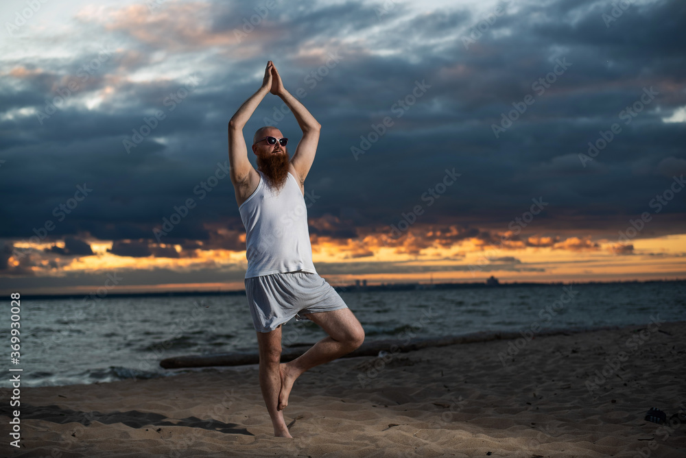 Obraz premium A bald man with a red beard practices yoga on the beach at sunset. A funny dude in a T-shirt and sunglasses meditates on the seashore.