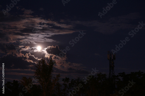 Color photography of an evening nature with a dark blue sky, a large full moon in the clouds. Planets Jupiter and Saturn. Trees and mobile tower. Beautiful moonlight.
