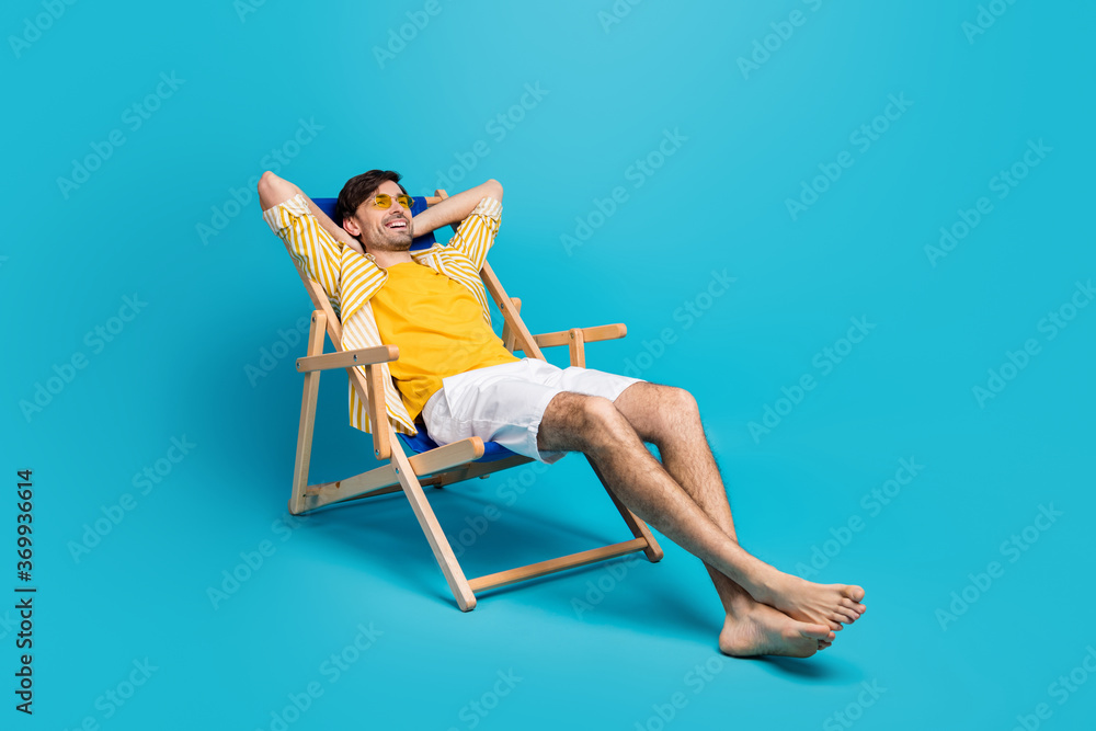 Full length profile side photo of positive guy traveler relax rest beach sun bathing lie comfort deckchair stretch hands wear white shorts isolated over blue color background - obrazy, fototapety, plakaty 