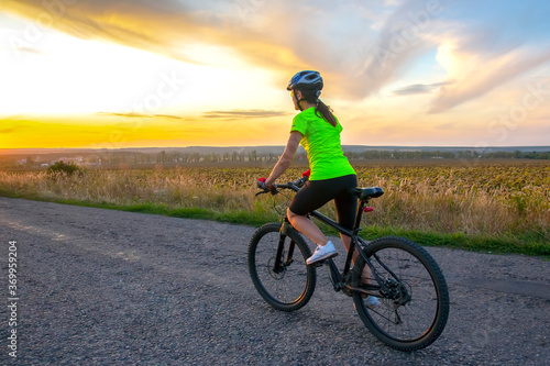 Fototapeta Naklejka Na Ścianę i Meble -  Beautiful girl cyclist riding a bike on the road towards the sunset. Nature and recreation. Hobbies and sports