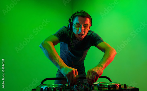 Young caucasian musician in headphones performing on green background in neon light. Concept of music, hobby, festival, entertainment, emotions. Joyful party host, DJ. Colorful portrait of artist.