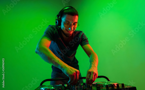 Young caucasian musician in headphones performing on green background in neon light. Concept of music, hobby, festival, entertainment, emotions. Joyful party host, DJ. Colorful portrait of artist.