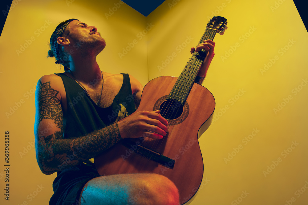 Obraz premium Young musician with guitar performing on yellow background in neon light. Concept of music, hobby, festival, entertainment, emotions. Joyful, inspired guitarist. Colorful portrait of artist.