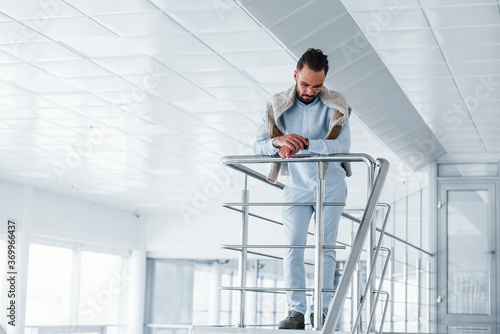 Leaning on silver colored railings. Young handsome man in formal clothes indoors in the office at daytime