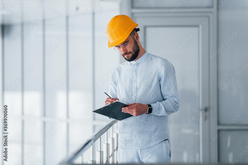Holding notepad. Engineer in white clothes and orange protective hard hat standing and working indoors Holding notepad. Engineer in white clothes and orange protective hard hat standing and working indoors