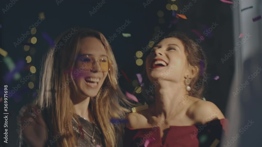 Close up couple of young multiracial girls wearing plush dresses enjoying glamorous party and having fun. Women dancing with falling confetti and sparkling or glowing sparklers or fireworks. Close up couple of young multiracial girls wearing plush dresses enjoying glamorous party and having fun. Women dancing with falling confetti and sparkling or glowing sparklers or fireworks.