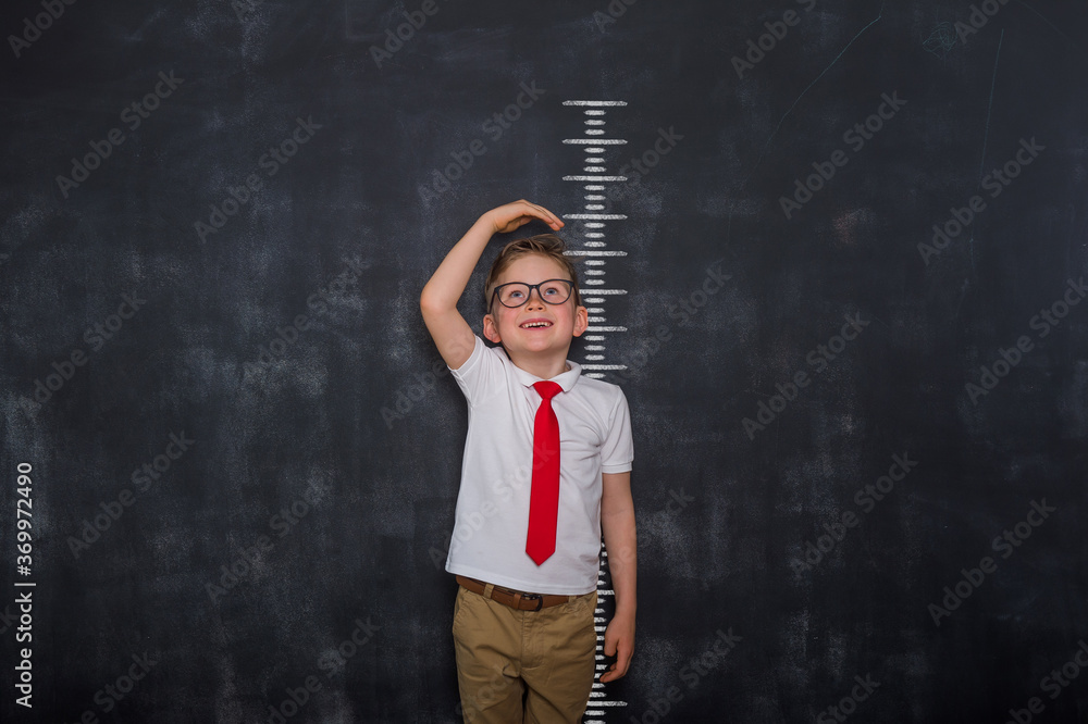 Little kid boy measuring himself. School boy measuring his growth in height against a blackboard scale. Back to school. Pupil in uniform - obrazy, fototapety, plakaty 