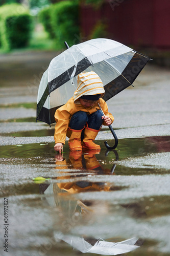 Fototapeta Naklejka Na Ścianę i Meble -  Kid in yellow waterproof cloak, boots and with umbrella playing outdoors after the rain