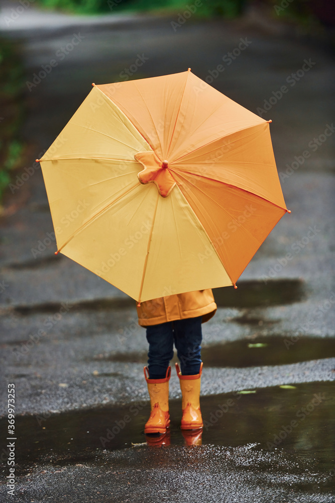 Obraz premium Kid in yellow waterproof cloak, boots and with umbrella playing outdoors after the rain