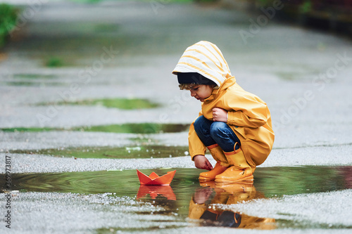 Fototapeta Naklejka Na Ścianę i Meble -  Kid in yellow waterproof cloak and boots playing with paper handmade boat toy outdoors after the rain