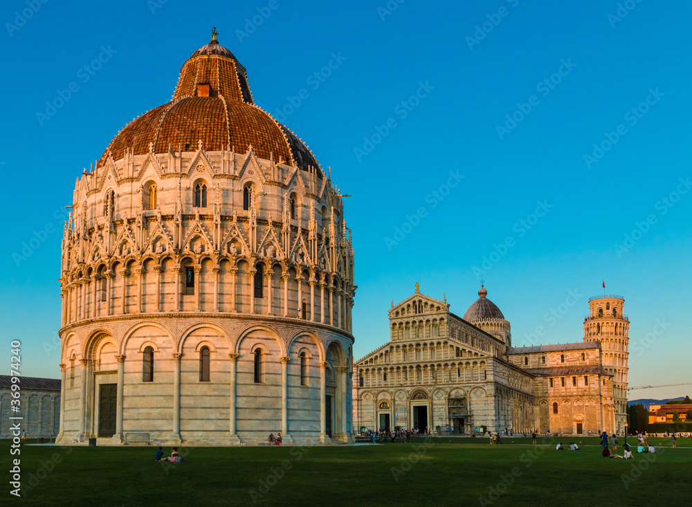 Picturesque view of the Pisa Baptistery of St. John in the foreground, the Duomo in the center, and the Leaning Tower in the background on the right, on a nice evening with a blue sky in Italy.  