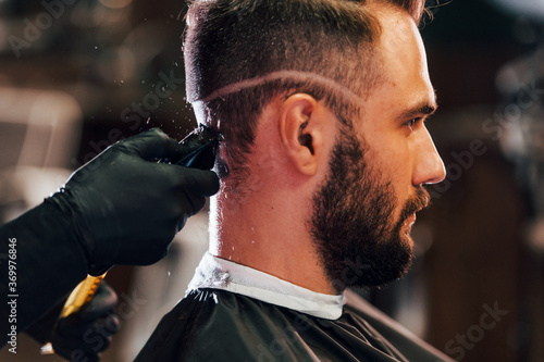 Young bearded man sitting and getting haircut in barber shop