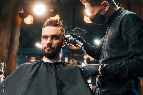 Front view of young bearded man that sitting and getting haircut in barber shop by guy in black protective mask