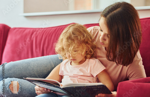 Reading big book. Young mother with her little daughter in casual clothes together indoors at home