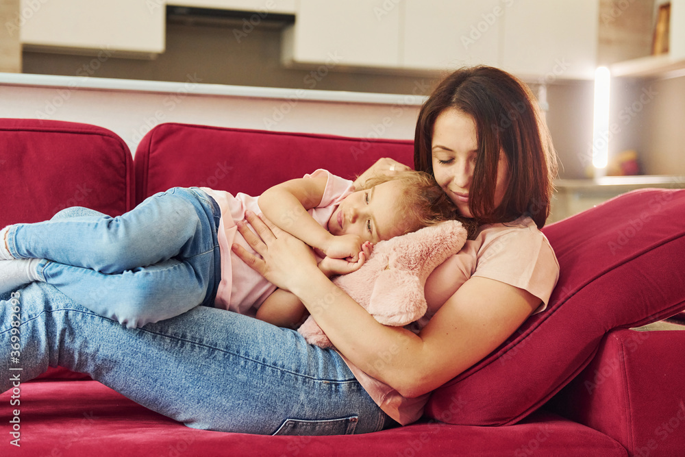 Lying down on red sofa. Young mother with her little daughter in casual clothes together indoors at home Lying down on red sofa. Young mother with her little daughter in casual clothes together indoors at home