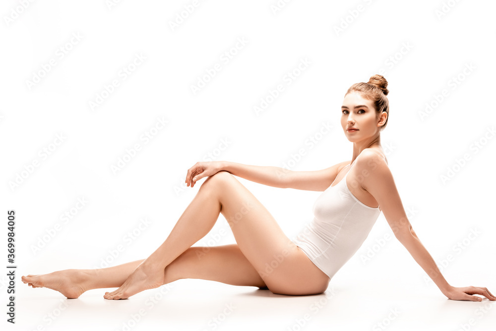 Fototapeta premium barefoot and slim young woman in bodysuit sitting on white