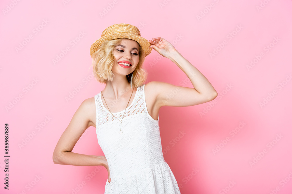 young woman touching straw hat and standing with hand on hip on pink young woman touching straw hat and standing with hand on hip on pink
