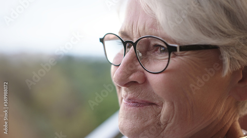 Senior gray haired woman with glasses standing alone on the balcony with smile but sad eyes. High quality 4k footage