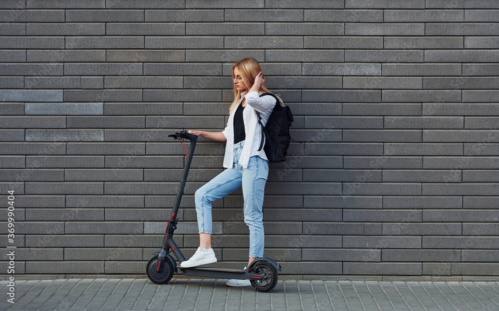 Side view of beautiful blonde in casual clothes that riding electric schooter outdoors at sunny daytime Side view of beautiful blonde in casual clothes that riding electric schooter outdoors at sunny daytime