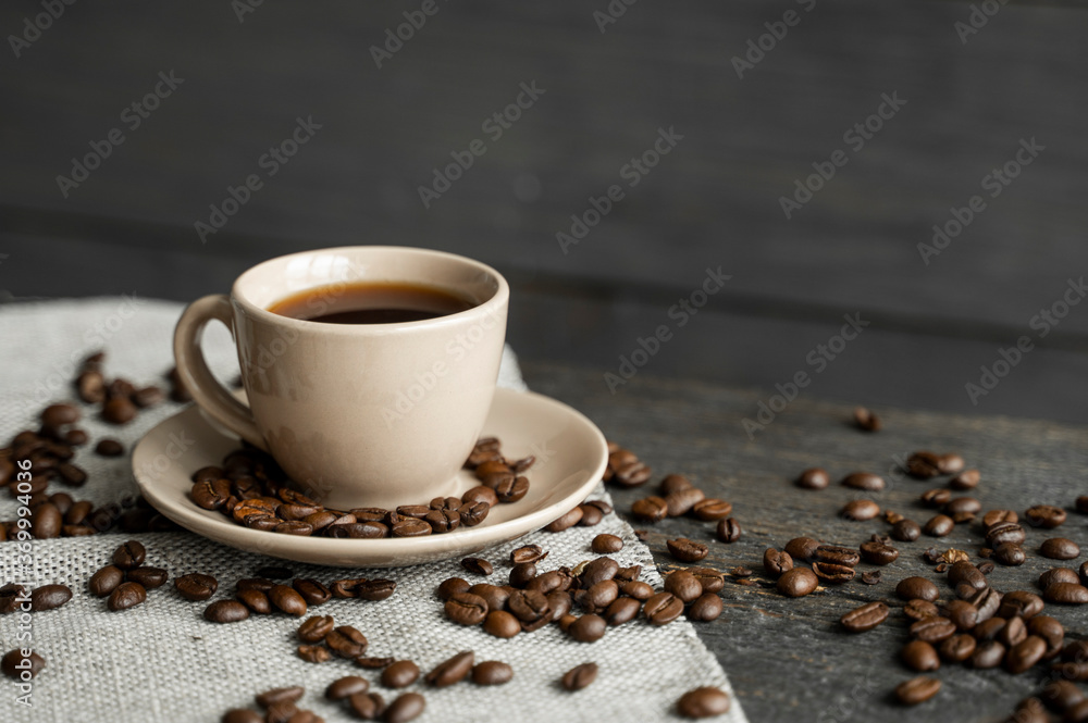 Obraz premium Coffee cup with roasted coffee beans on wooden table background. Mug of black coffe with scattered coffee beans on a wooden table. Fresh coffee beans.