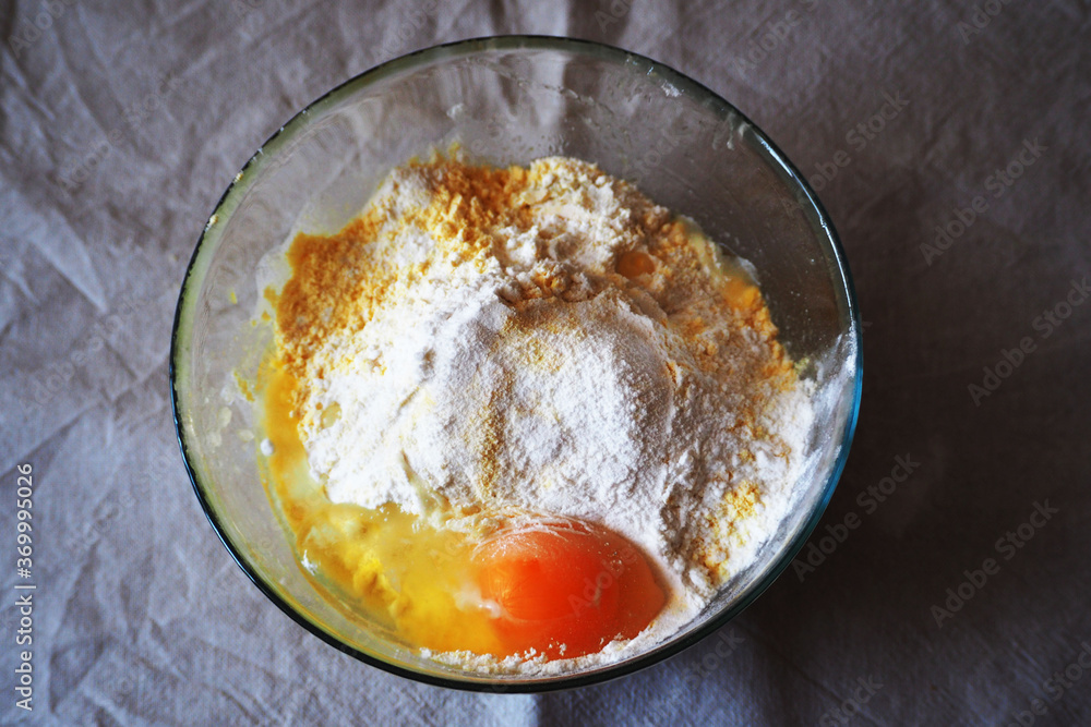 transparent glass round bowl with corn and rice flour with one egg on beige tablecloth top view. Ingredients before baking gluten-free at home transparent glass round bowl with corn and rice flour with one egg on beige tablecloth top view. Ingredients before baking gluten-free at home