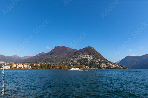 Stunning panorama view of Lugano Lake, cityscape of Lugano, mountain Monte Bre and Swiss Alps on a sunny autumn day with blue sky cloud, cruise ship in foreground, Canton of Ticino, Switzerland