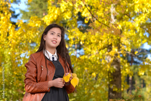 A girl in a brown leather jacket holds yellow leaves. Portrait of a beautiful young girl with long black hair against the background of the autumn forest.
