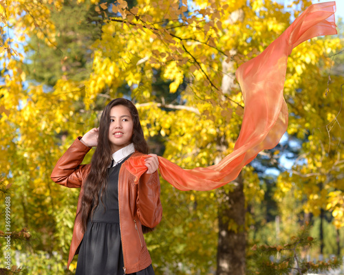 A girl in a brown leather jacket holds an orange silk scarf. Portrait of a beautiful young girl against the background of the autumn forest with a developing orange scarf in her hand.