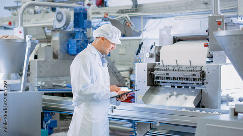 Young Male Quality Supervisor or Food Technician is Inspecting the Automated Production at a Dumpling Food Factory. Employee Uses a Tablet Computer for Work. He Wears White Sanitary Hat and Work Robe.