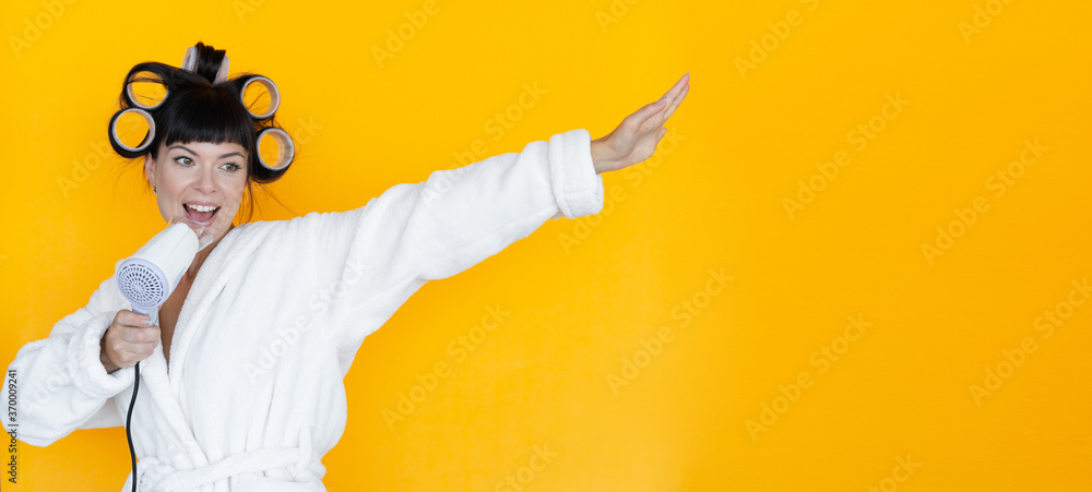 young beautiful woman housewife in a white robe. curlers on the head. Yellow background. Glove-shaped washcloth. hold the hair dryer in your hands