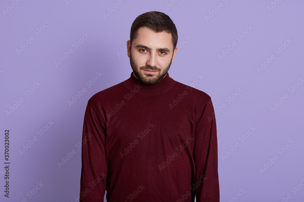 Young unshaved man wearing burgundy sweater posing against purple background with serious facial expression, looking directly at camera with confident look. Young unshaved man wearing burgundy sweater posing against purple background with serious facial expression, looking directly at camera with confident look.