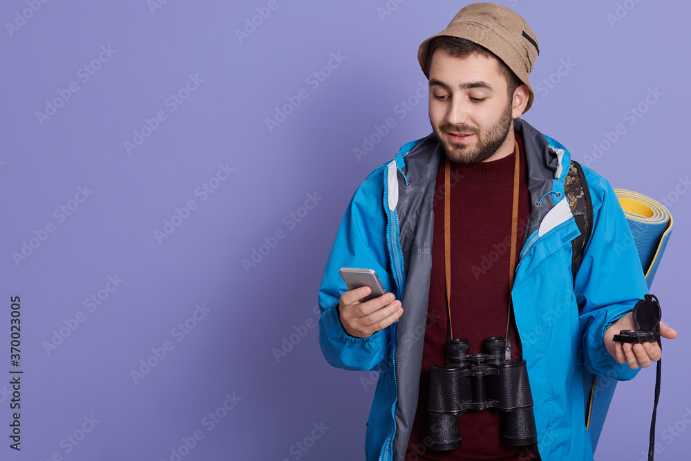 Attractive man tourist backpacker using internet application on modern smart phone, posing with mat, binocular and backpack against blue background. Attractive man tourist backpacker using internet application on modern smart phone, posing with mat, binocular and backpack against blue background.