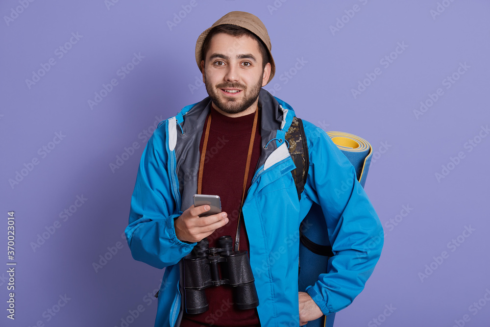 Smiling traveler man in cap and with backpack isolated on blue background. Tourist traveling on weekend getaway, being in good mood, holding smart phone. Tourism, discovering, hiking. Smiling traveler man in cap and with backpack isolated on blue background. Tourist traveling on weekend getaway, being in good mood, holding smart phone. Tourism, discovering, hiking.