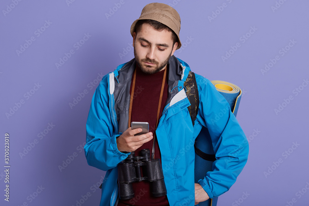 Young bearded man sending messages for his family from his cellphone, during hiking. Traveler in hat and jacket using mobile phone application, has binocular and mat. Young bearded man sending messages for his family from his cellphone, during hiking. Traveler in hat and jacket using mobile phone application, has binocular and mat.