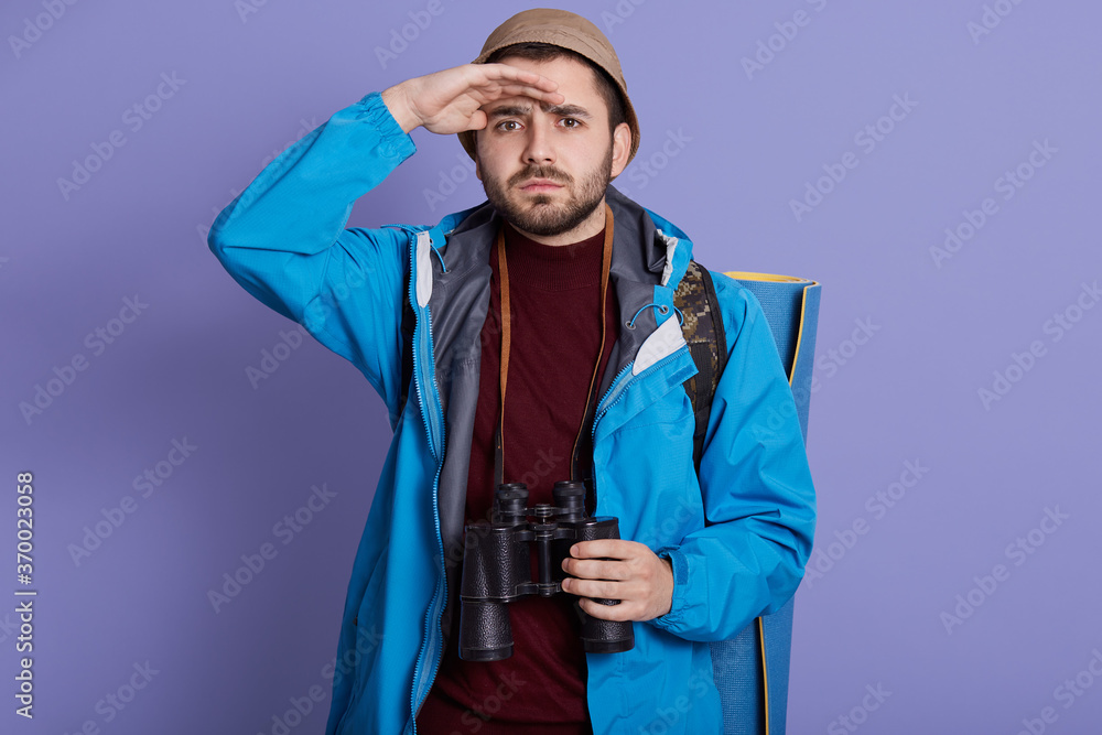 Guy tourist hiker with backpack and travel mat, wearing jacket and hat, stands with binocular over his neck isolated over blue background, keeps palm near forehead and look in distance. Guy tourist hiker with backpack and travel mat, wearing jacket and hat, stands with binocular over his neck isolated over blue background, keeps palm near forehead and look in distance.