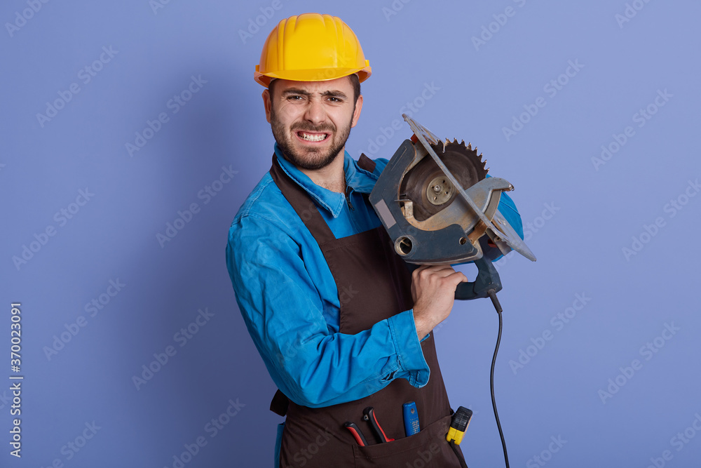 Carpenter working with circular saw looking directly at camera wit angry facial expression, wearing yellow helmet and brown apron, standing against blue background. Carpenter working with circular saw looking directly at camera wit angry facial expression, wearing yellow helmet and brown apron, standing against blue background.