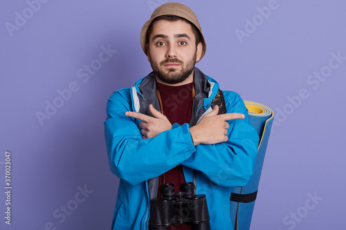 Young Caucasian traveler backpacker man isolated over blue background, pointing both sides, dos not know where to go, wearing jacket, hat, posing with mat, backpack and binocular. © sementsova321
