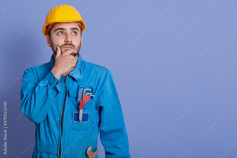 Pensive guy wearing yellow helmet and blue overalls, bared boy with wrench, looking aside with thoughtful facial expression. Copy space for advertisement. Pensive guy wearing yellow helmet and blue overalls, bared boy with wrench, looking aside with thoughtful facial expression. Copy space for advertisement.
