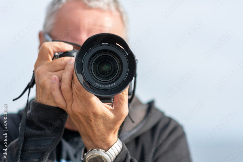 A middle-aged man, in his 50th, takes a picture with a modern mirrorless camera. The camera covers his face. Blue hazy sky in the background