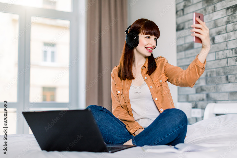 Fototapeta premium Distance learning, online education and work. Young happy smiling woman in headphones having a video cal, while working from home on bed using computer and mobile phone