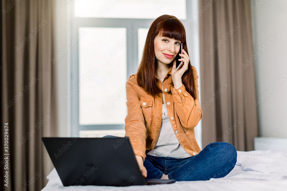 Pretty young business woman in casual wear, sitting on bed in lotus pose, working with laptop computer on a bed, having a call with coworkers or clients. Work from home concept Pretty young business woman in casual wear, sitting on bed in lotus pose, working with laptop computer on a bed, having a call with coworkers or clients. Work from home concept