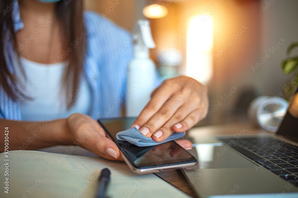 Fototapeta premium Woman wiping mobile phone while working from home. Woman cleaning her phone with disinfectant. Young woman wiping her electronic equipment with alcohol wet wipe